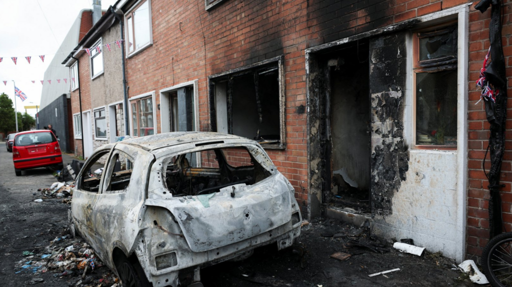 The remains of a burned out white car on the kerb outside a row of red brick houses in Belfast. The house nearest to the car also shows clear signs of fire damage, with black burn stains around windows and the front door