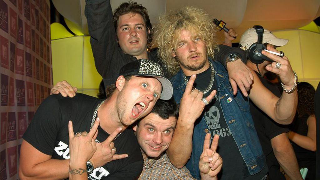 Photograph of Dirty Sanchez stars. Mathew Pritchard (right) stands holding a cigarette in his left hand, and poses with rock fingers in his right hand. He has blonde big hair and looks dazed at the camera. He wears a denim vest and a black top with a silver chain.  Lee Dainton ( left) holds two rock fingers using his hands and poses with his mouth stretched wide open. He wears a silver chain and a black short sleeve t-shirt and has blue eyes. Dan Joyce stands behind the group. He has brown long hair cut into a mullet and brown eyes. He has one arm in the arm and own arm around Pritchard. Mike "Pancho" Locke pops his head in the centre of the group and poses with a peace sign.