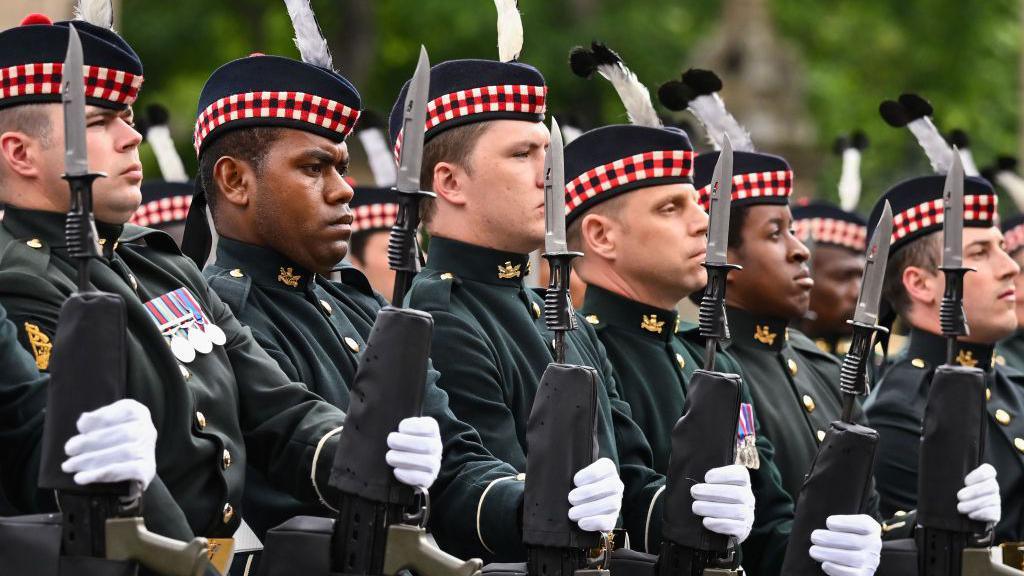 Eight soldiers from the Royal Regiment of Scotland standing in a line in dark ceremonial uniforms. They are all wearing white gloves and holding rifles.