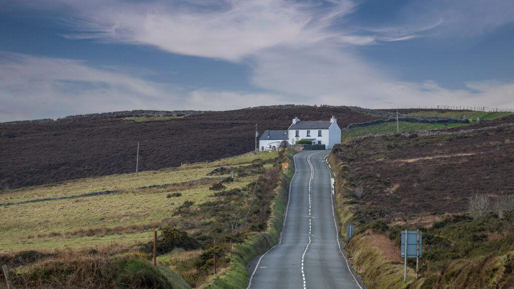 The Mountain Road with a white cottage at the end of the road and greenery around the road.