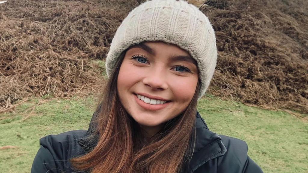 A young woman smiles broadly at the camera. She is stood outside with grass and moorland behind her. She wears a black coat and a beige knitted hat. She has medium-length brown hair and is wearing natural make-up.