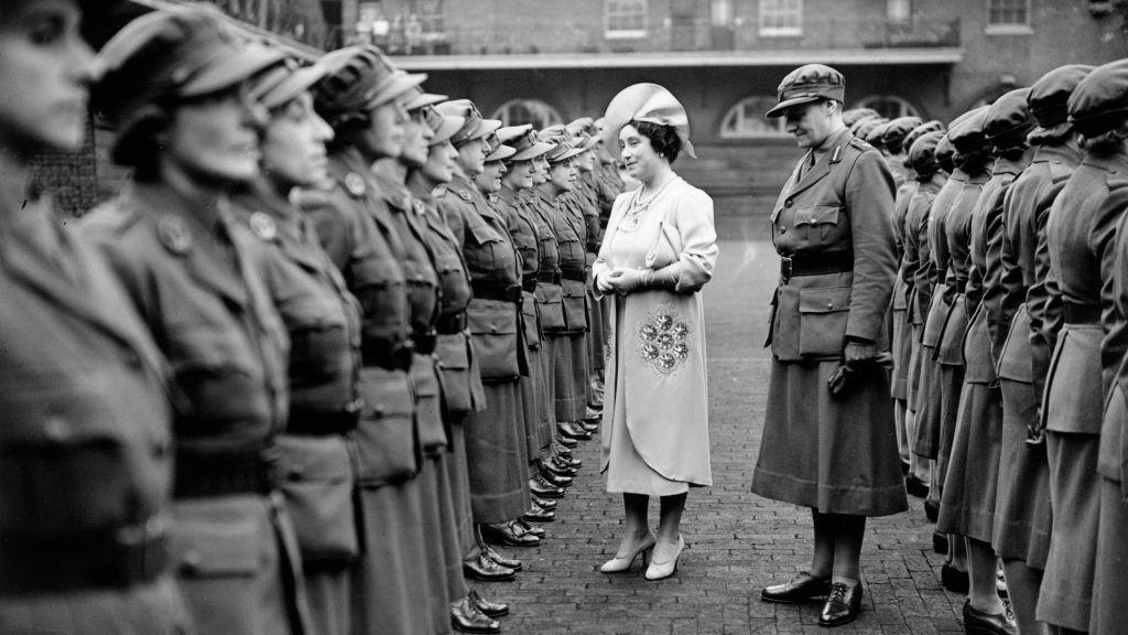 Women in World War Two military uniform line up for inspection by the Queen Consort who is wearing an elaborate hat. 