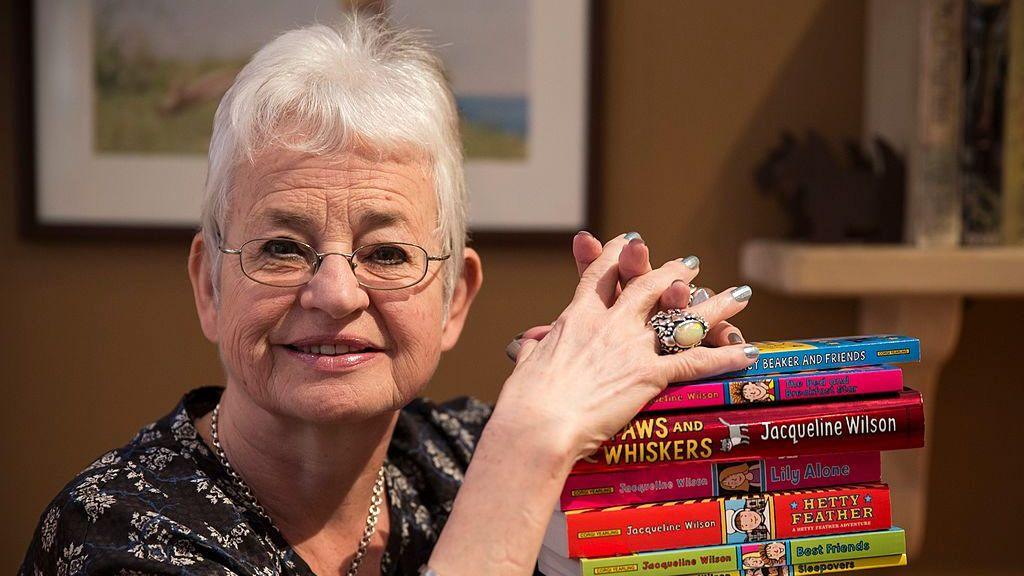 Dame Jacqueline Wilson looks at the camera, holding onto a pile of her books.