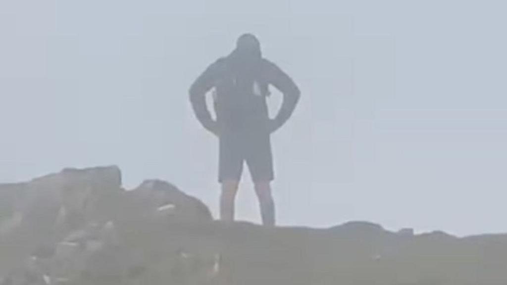 A grainy photograph of John Hall from behind with his hands on his hip, he's at the peak of Helvellyn in the Lake District. 