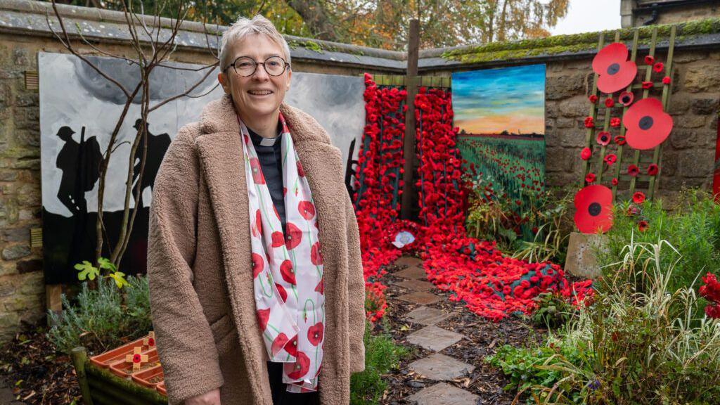 Rev Paula Challen standing in a beige coat, poppy scarf and dog collar, in the Remembrance Garden.