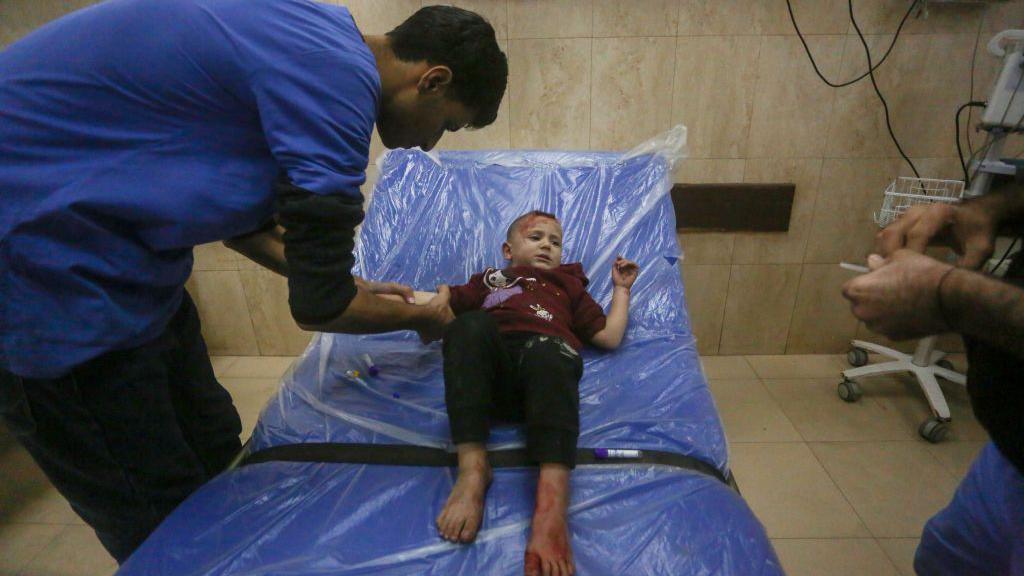 An injured child with bare feet lies on a blue bed covered with transparent plastic sheeting, while a doctor dressed in a blue overall bends over him and holds his right arm.