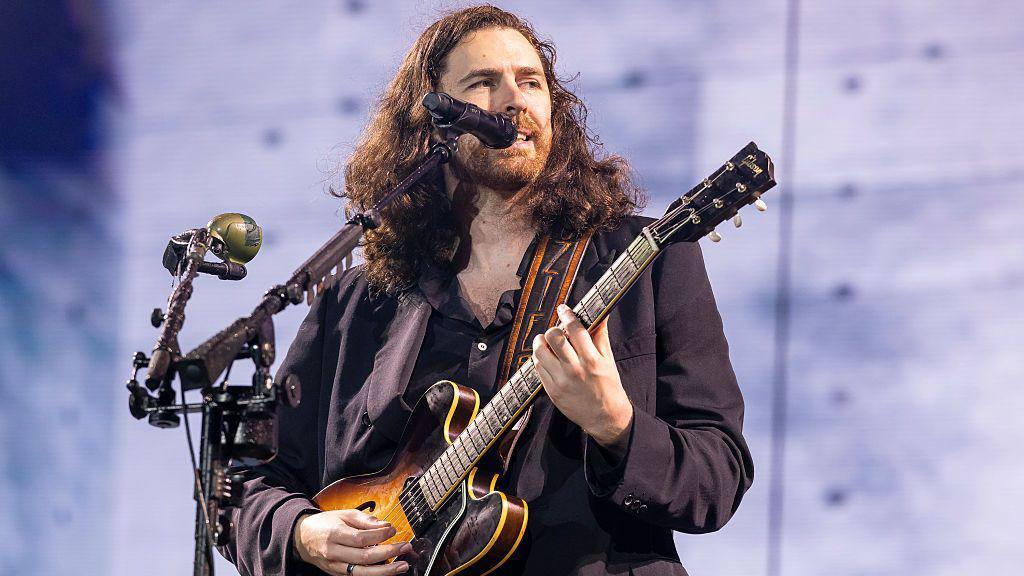 A man with shoulder-length brown wavy hair plays guitar on stage.