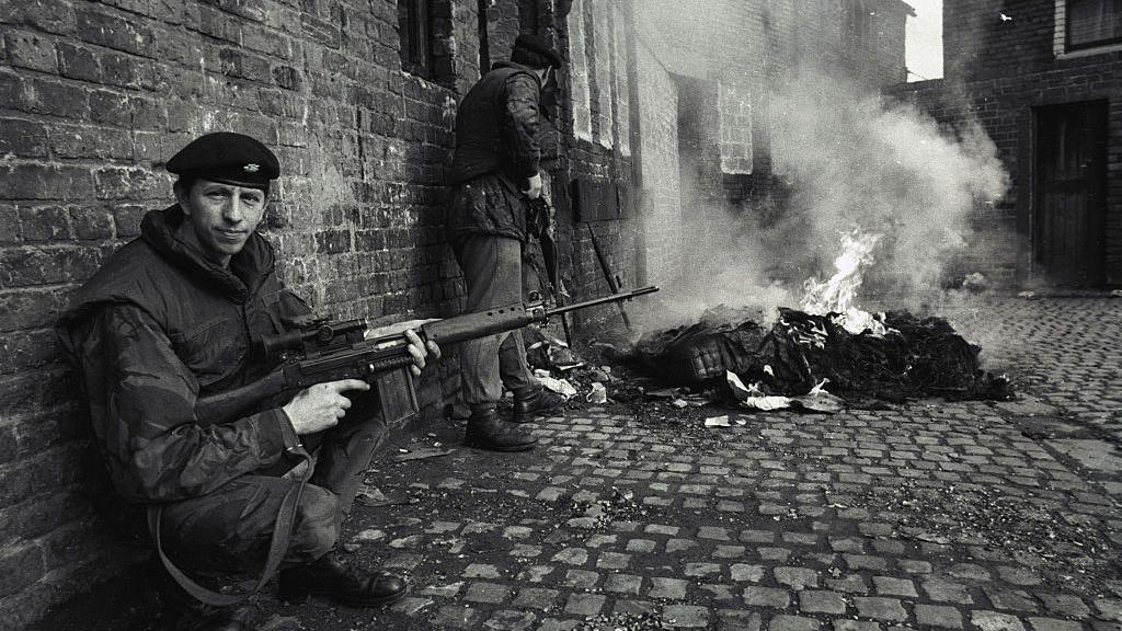 A black and white photo shows a solider in army uniform holding a rifle, crouch with his back towards a brick wall, he looks at the camera. Behind him to his left is a solider with his back to the man crouching, he is also in uniform holding a rifle. There is a pile of debris on fire to the right of the man standing.