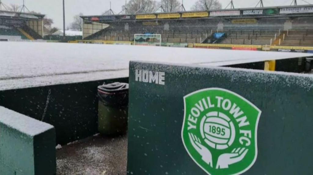 Snow covering Yeovil Town Football Club pitch and stands. In the foreground is the club's green crest which says its name and has an image of a football and two hands underneath.