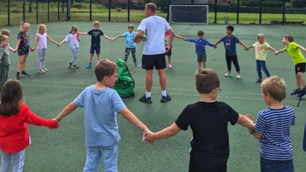 Children taking part in the Healthy Me camp. They stand in a circle holding hands on an outdoor court, with a male teacher in the centre.