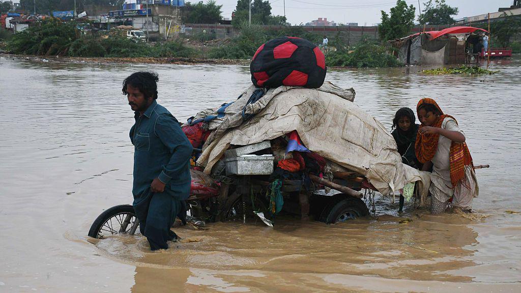 A man towing his pile of belongings on a motorbike through knee-deep brown floodwater. As he drags the bike in the front, two women are wading behind the bike in the back.