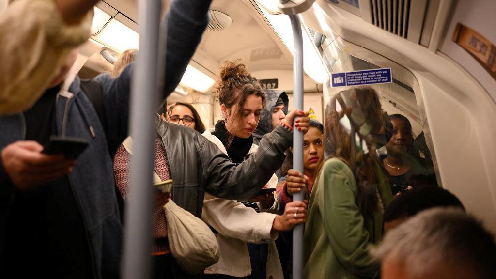 Interior shot of crowded Underground train, with people wearing coats and looking at phones.