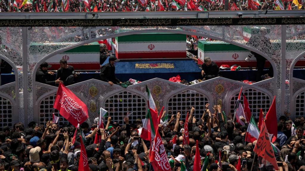 Coffins, painted with the Iran flag, sit in truck with see-through sides. A huge crowd of people are in front and behind the vehicle, with people waving flags and raising their hands in the air.