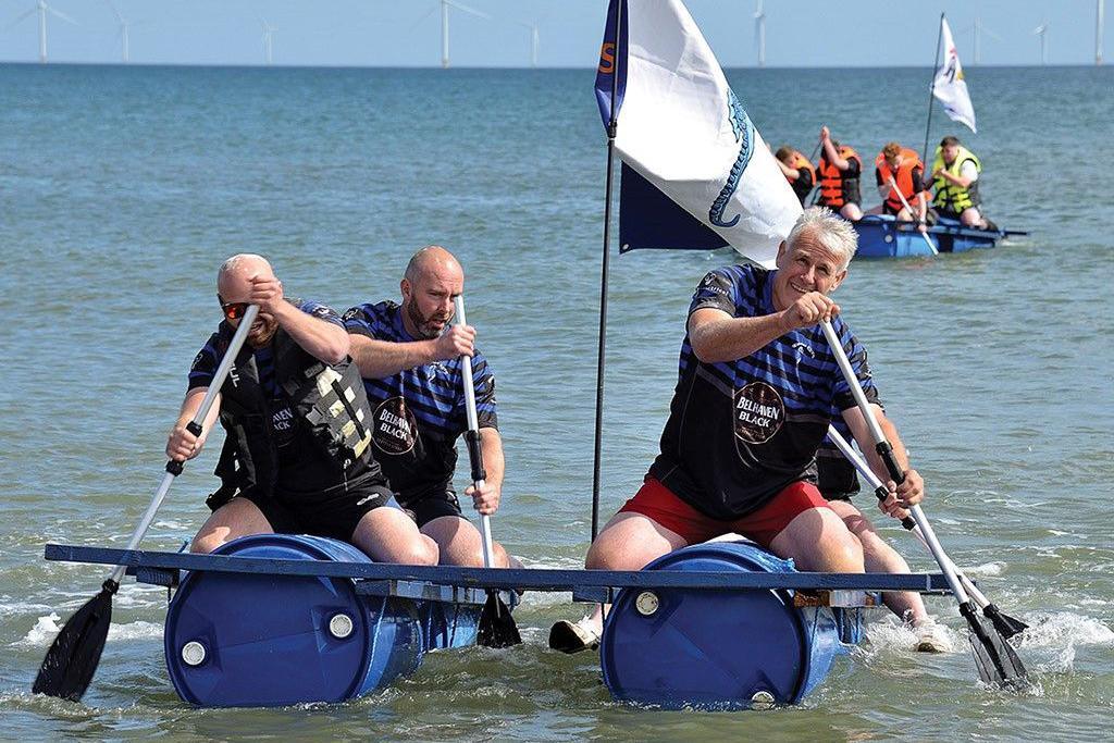 Three men with life jackets on a raft made of out barrels in the sea. There is another group on a similar raft in the background.