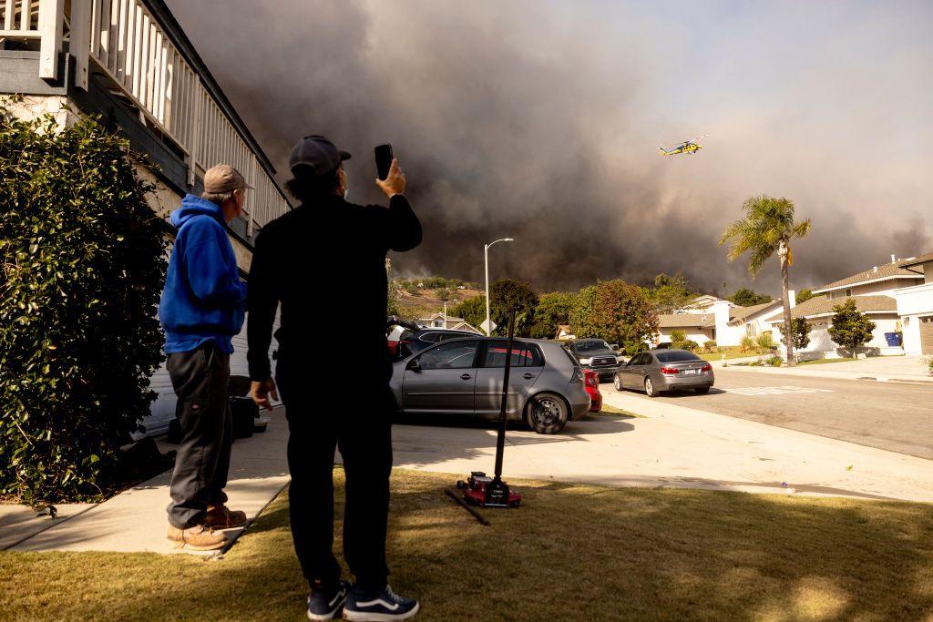 Two men, one wearing a blue hoodie and beige cap and another wearing a black top and black cap holding up a camera phone, watch a firefighting helicopter fly above a plume of smoke