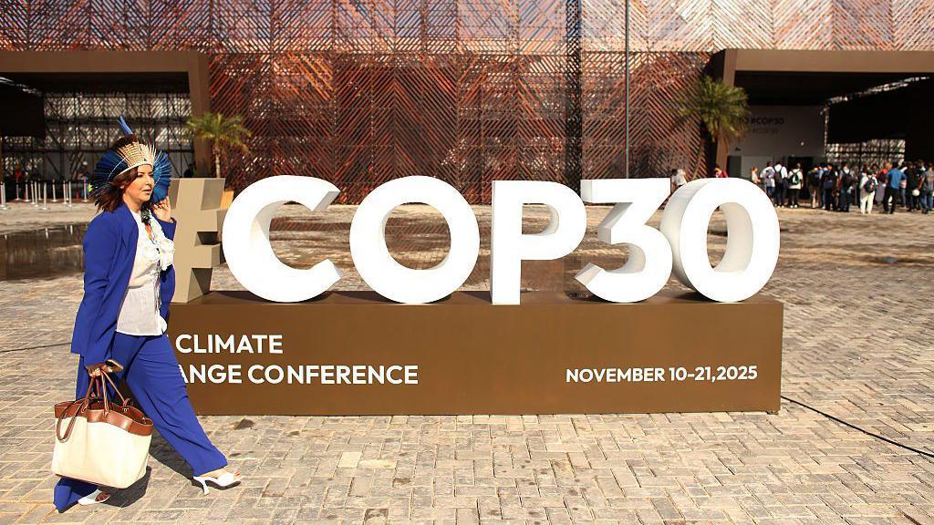 A woman in a blue suit and traditional headress, carrying a large white handbag, walks past a large three-dimensional sign advertising "#COP30 climate change conference"