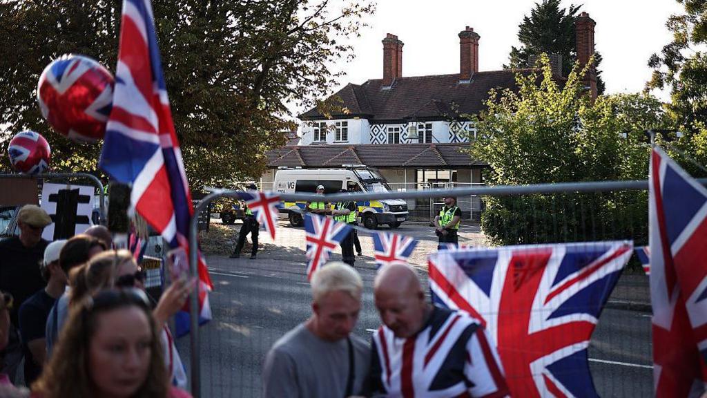 A picture of the Bell Hotel in Epping. There is a police van and four officers outside the hotel. In the foreground there is a metal fence on which protesters have hung Union Jack bunting and flags. In front of the fence, there are seven people. Some are holding Union Jack balloons. 
