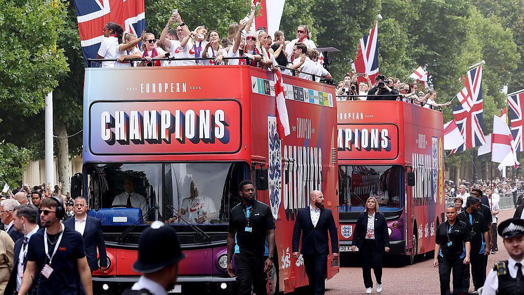 Two buses make their way down The Mall with members of staff walking alongside.