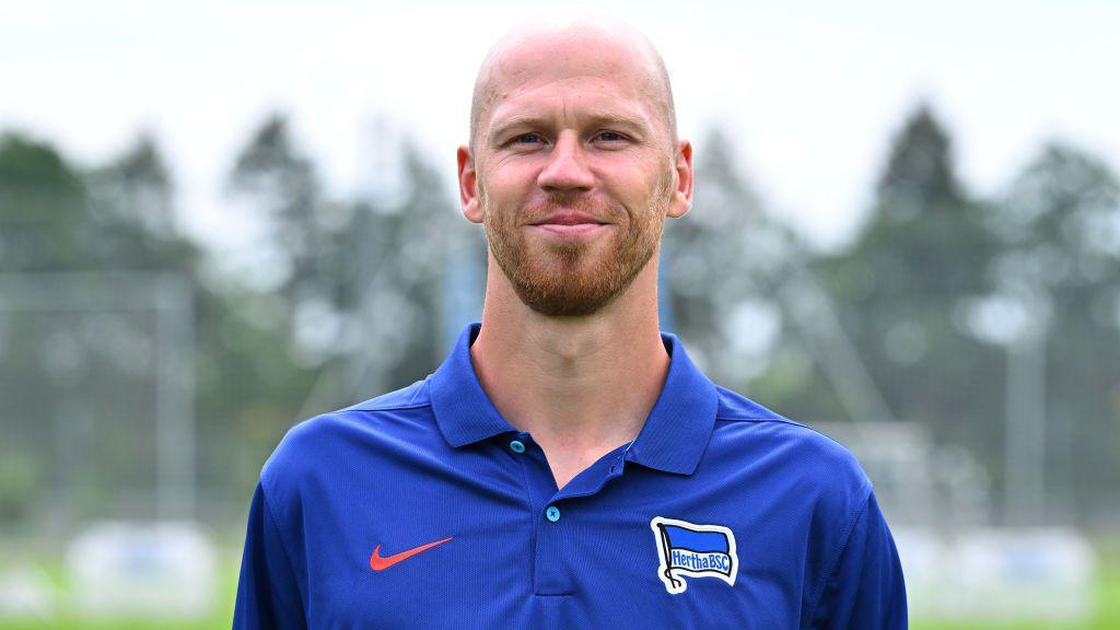 A bearded, shaven headed Gabor Ruhr faces the camera wearing a blue Hertha Berlin polo shirt