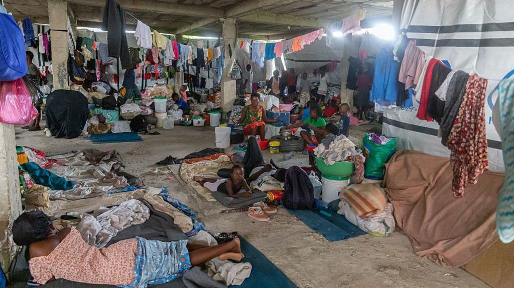 People in a makeshift shelter in Port-au-Prince, sitting and lying on mats on a concrete floor, with lines of washing strung above them. Several water buckets and piles of possessions can be seen in the background.
