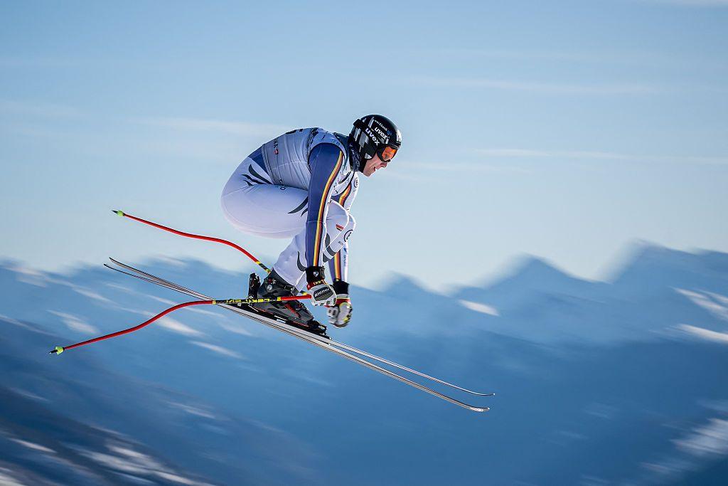 German skier Luis Vogt launches into the air during a high-speed downhill run, body tucked tight, skis angled forward, with blurred mountain peaks behind.