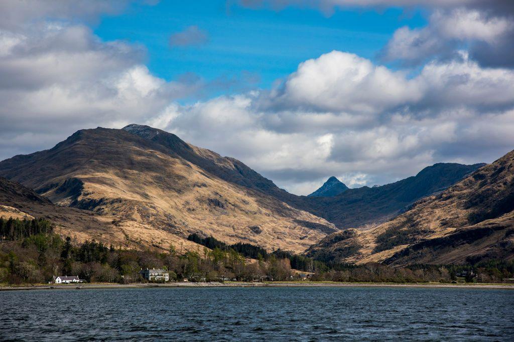 The village of Inverie on the Knoydart Peninsula. There are a handful of houses on the banks of a loch, with mountains and blue skies in the background.