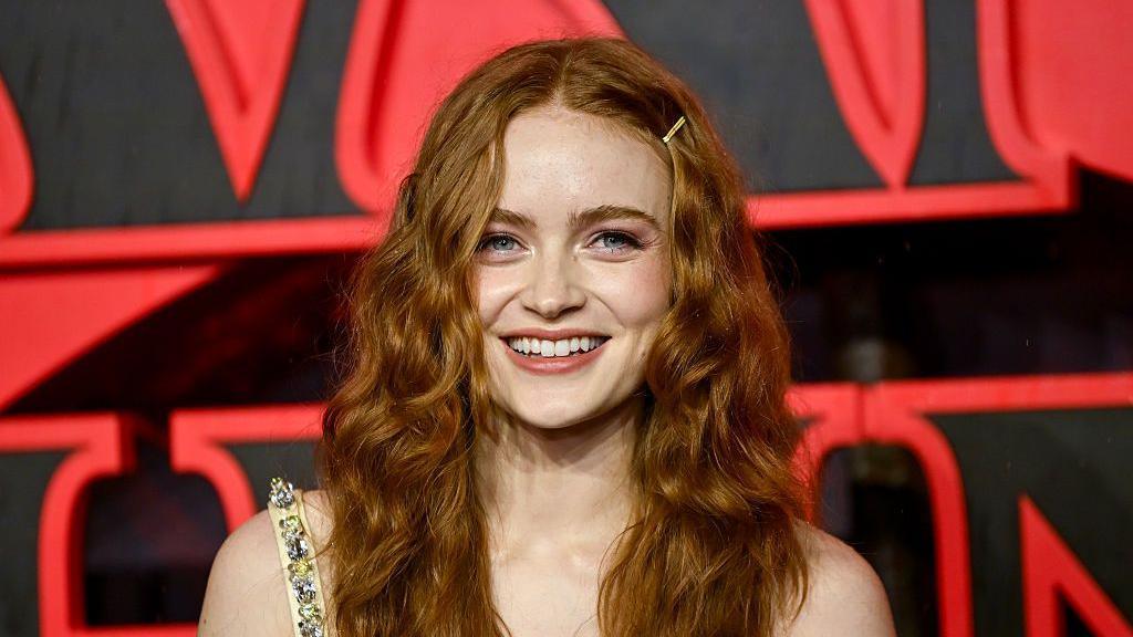 Sadie Sink is smiling close up to the camera, she has red wavy hair to her shoulders. She has blue eyes and a gold clip in her hair. Behind her is the Stranger Things logo. 