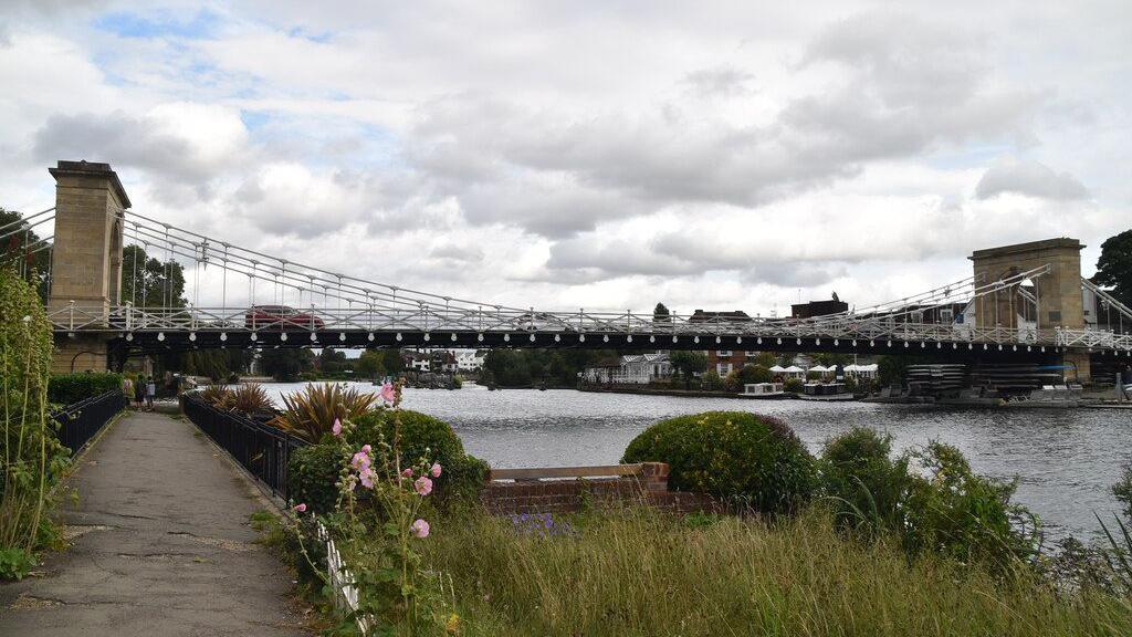 The Grade I listed Marlow Bridge, showing a bridge over the Thames, with flowers and foliage on the bank, cars crossing the bridge and water underneath it. There are building to the right. There are two bridge structures on the bridge.