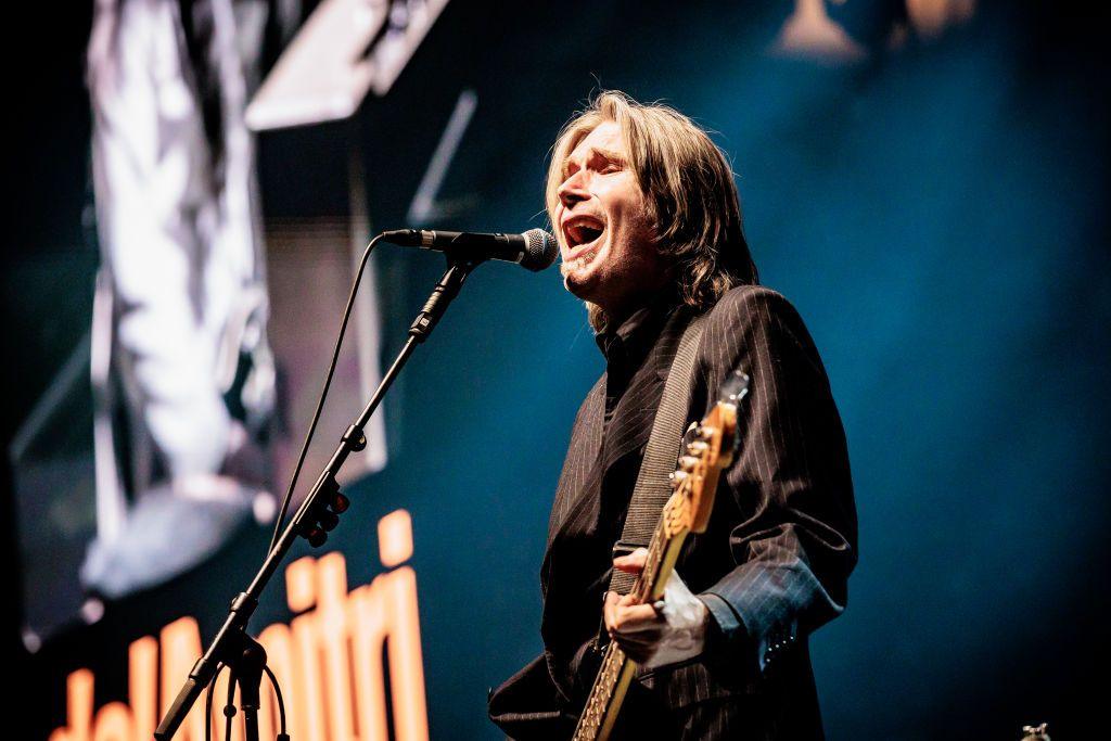 Justin Currie is holding a guitar and singing into a microphone.