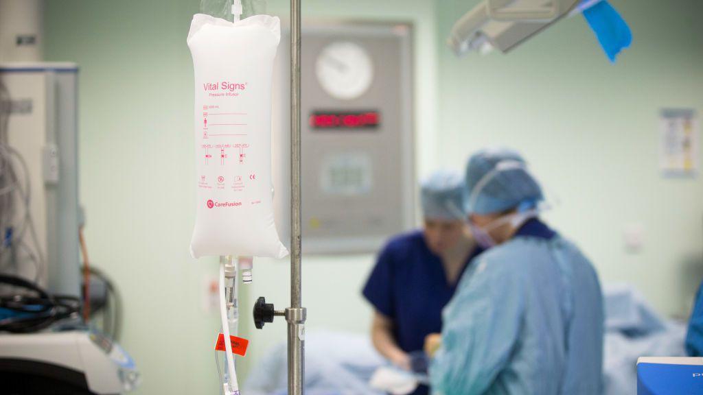 A close up of an IV drip in a hospital surgery. There are two medical professionals in scrubs in the background