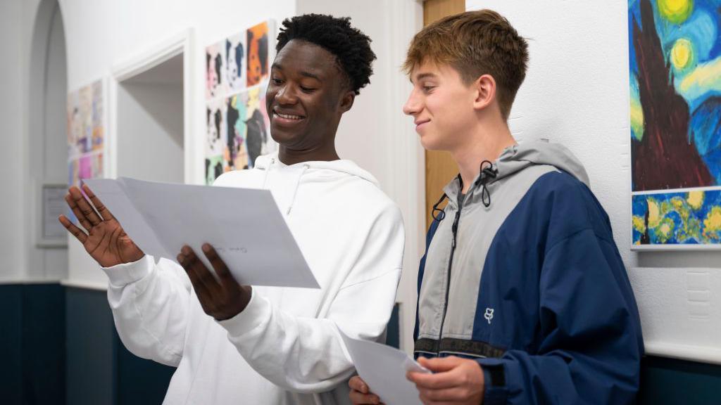 Two students smile and hold their GCSE results in school