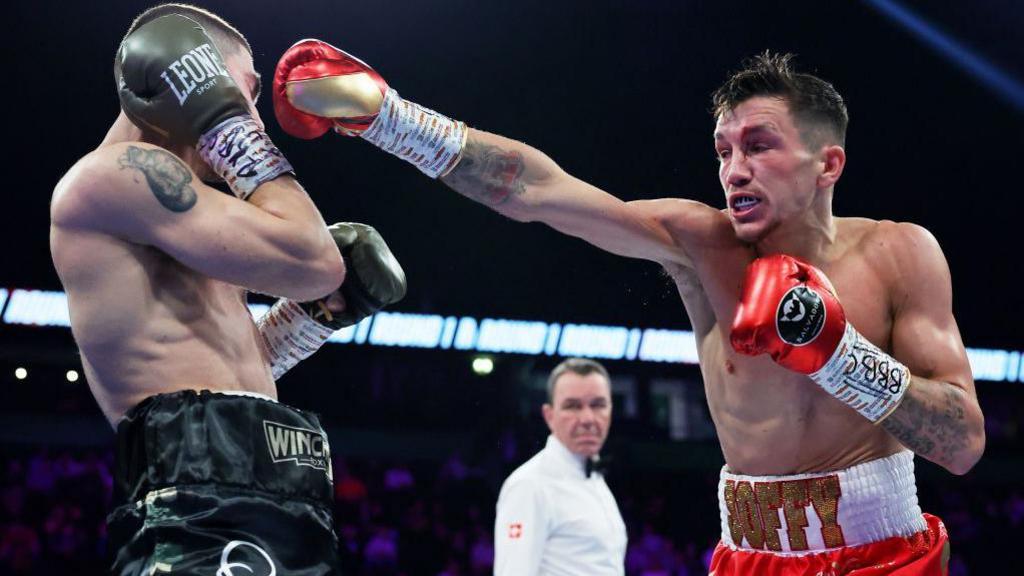 Davies and another boxer are shirtless in a boxing ring. There is a referee blurred in the background. Davies has his arm outstretched in a punch, wearing red boxing gloves. The other boxer has his hands shielding his face and is wearing black shorts and boxing gloves.