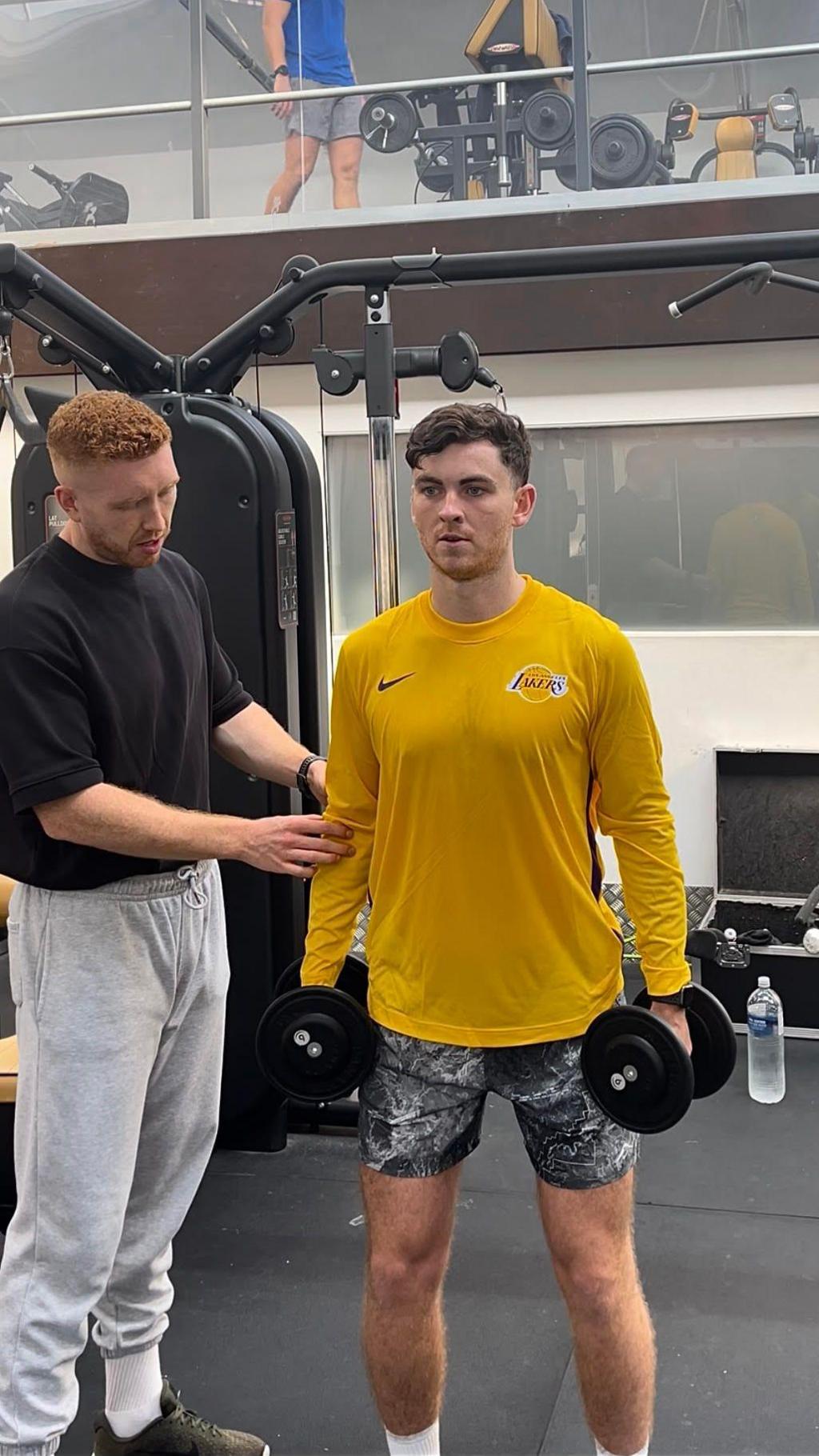 Two men standing in a gym, one is wearing a yellow top and black/white patterned shorts, holding a dumbell in each hand, and is slightly leaning forward. Sean is stood to his right touching his arm, and Sean is wearing a black top and light grey joggers, white socks and trainers.