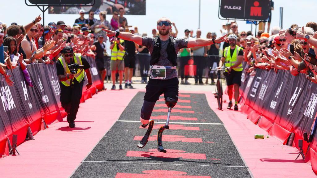 A male double amputee crosses the finish line of a race with his arms stretched wide as people cheer and applaud. 