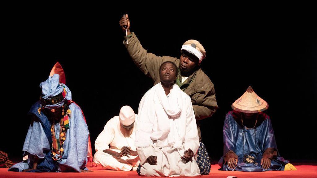 Five performers kneel on stage during the performance. The actor in the centre wears white as a man behind him in green points a dagger to his throat. Either side of them are two actors in blue - Saturday 8 March 2025.