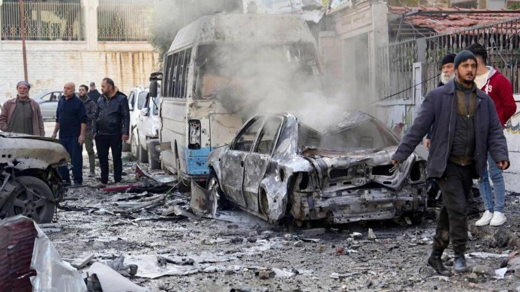 People survey the damage after an air strike in Idlib, Syria. A car is seen smoking and people in jackets walk around to survey the damage.