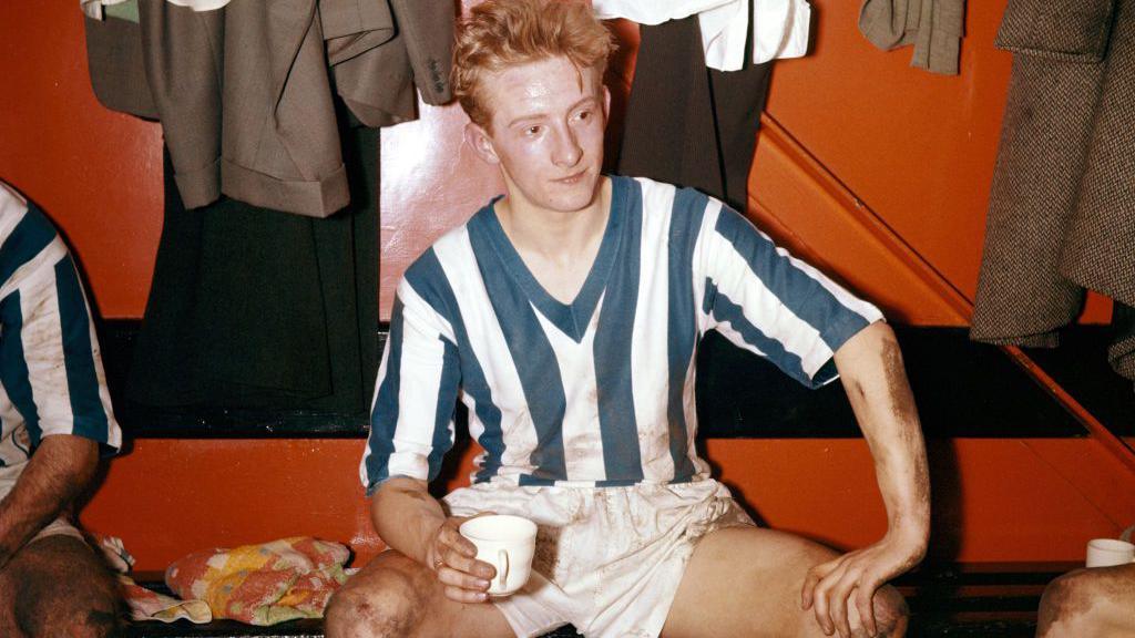 Denis Law drinks a cup of team after a Huddersfield Town game in 1957 when he was 16. He wears a blue and white striped football strip with white shorts. He appears to be sitting in the changing room, with clothes hanging on pegs behind him. 