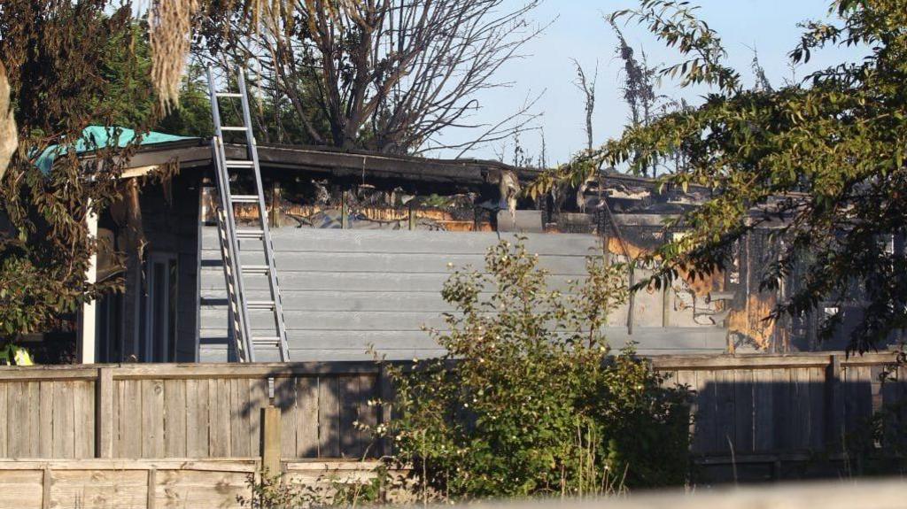 The top of a burnt outbuilding is visible. There is a ladder leaning against the building and there are trees around it.