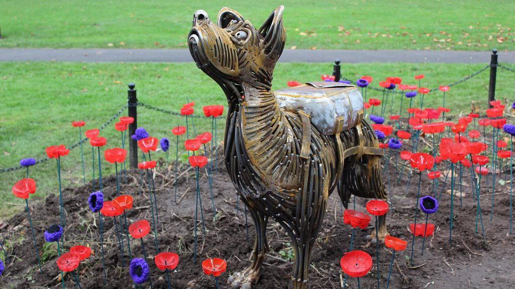 A metal sculpture of Bing, an Alsatian and Collie cross, from Loughborough, Leicestershire, surrounded with poppies in a flower bed in Queen's Park, in Loughborough.