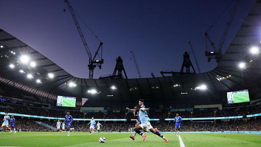 Jeremy Doku beats a Leicester defender at the Etihad Stadium with cranes in the background