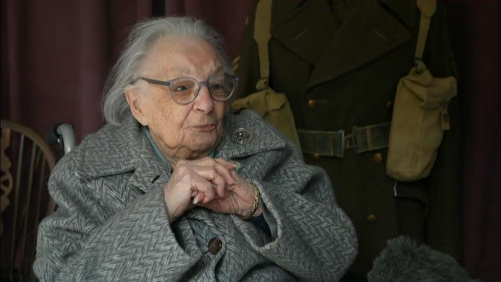 A woman wearing a grey coat sits with her hands clasped together.