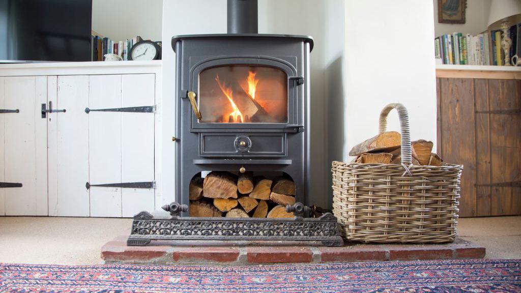 A log burner with a fire inside, and a stack of logs underneath with a basket next to it. There's a red and blue carpet in front of it and cupboards on either side.