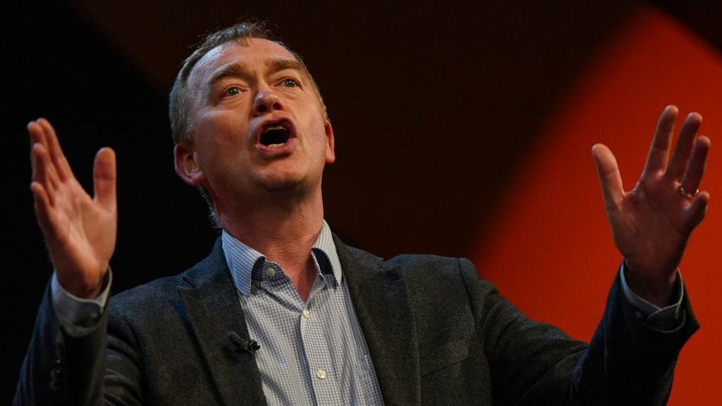 Tim Farron speaking at the 2024 Liberal Democrat conference. He is using his hands to emphasise his remarks. He is wearing an open-necked shirt with a check pattern and a grey blazer.