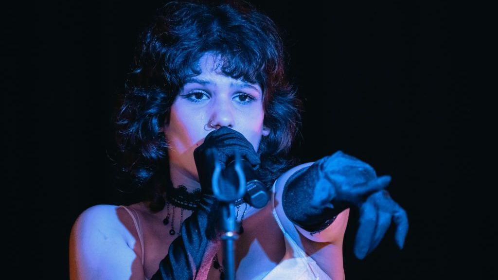 A singer with black hair styled in a short, 1920s-style bob sings into a microphone. She's wearing elbow-length, black velvet gloves. One hand holds the microphone and her free hand is pointing off-camera at someone or something in front of her.