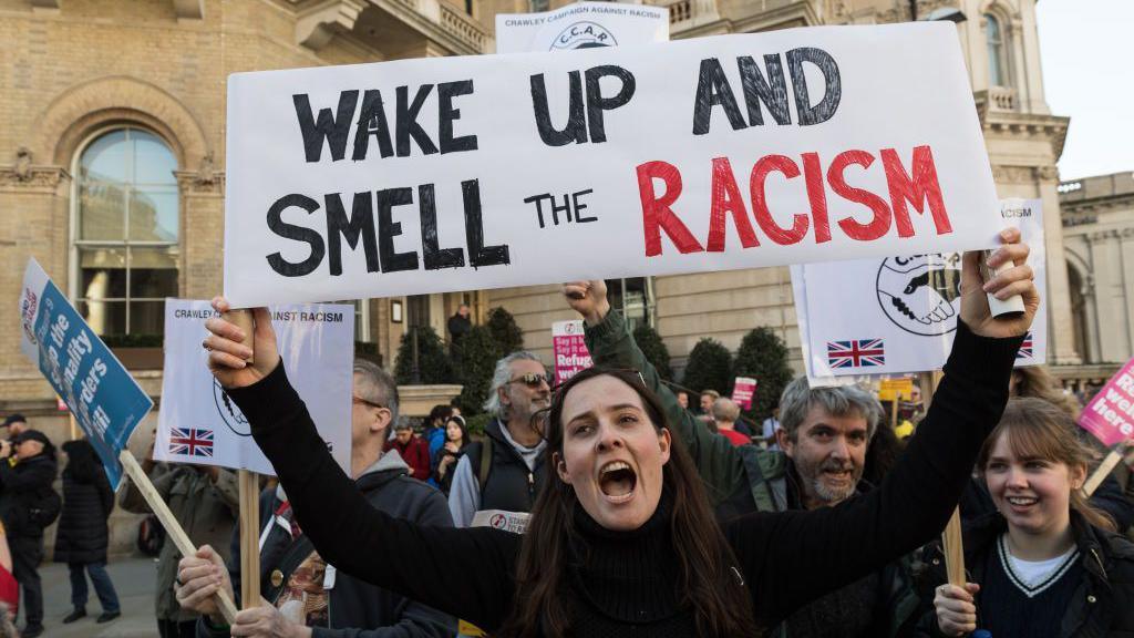 Protestors in Hackney including one prominently holding a banner stating: Wake up and smell the racism