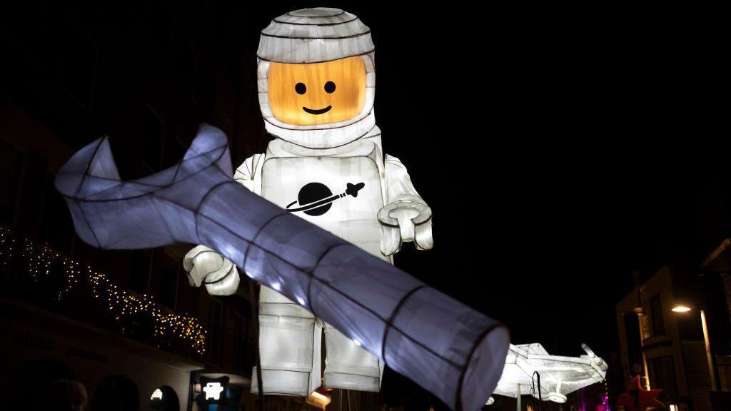 A large lantern in the shape of a lego astronaut is seen in front of a dark sky. There is also a lantern shaped like a wrench held in front of it and what looks like a smaller spaceship in the background, all illuminated from the inside.