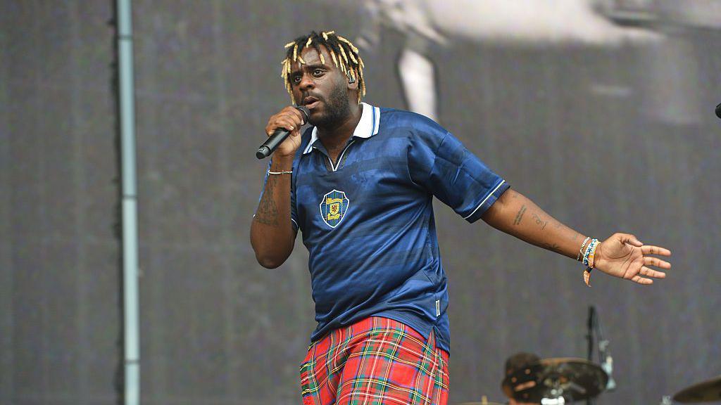 Myles Smith sings onstage while wearing a Scotland football top and a kilt