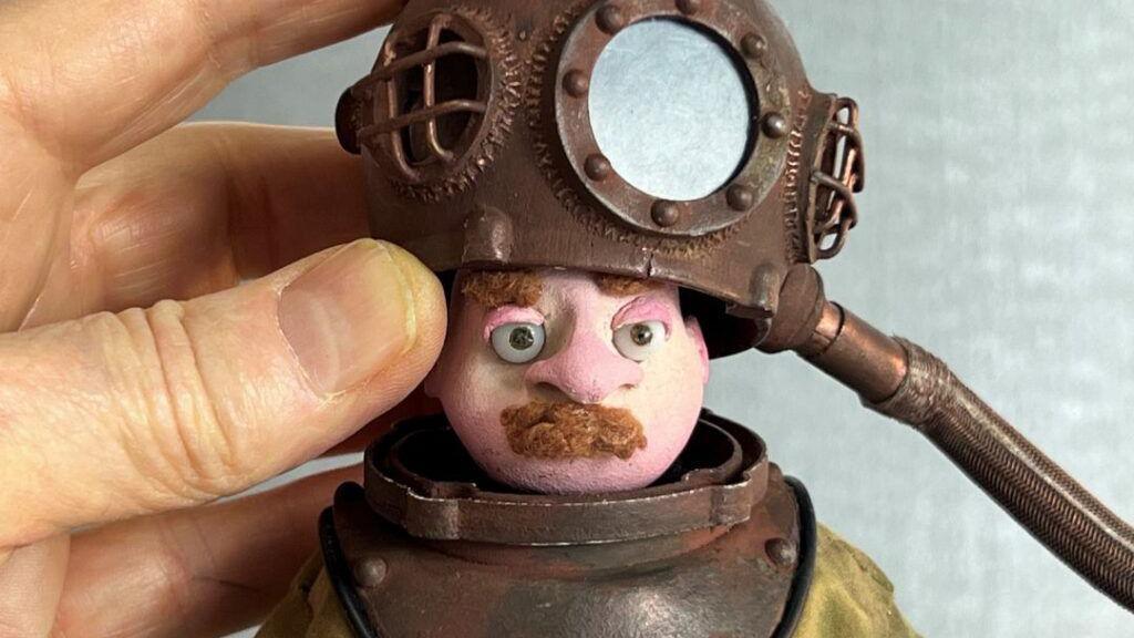 A comparatively large hand is holding a figure of a moustached man dressed as a deep sea diver.