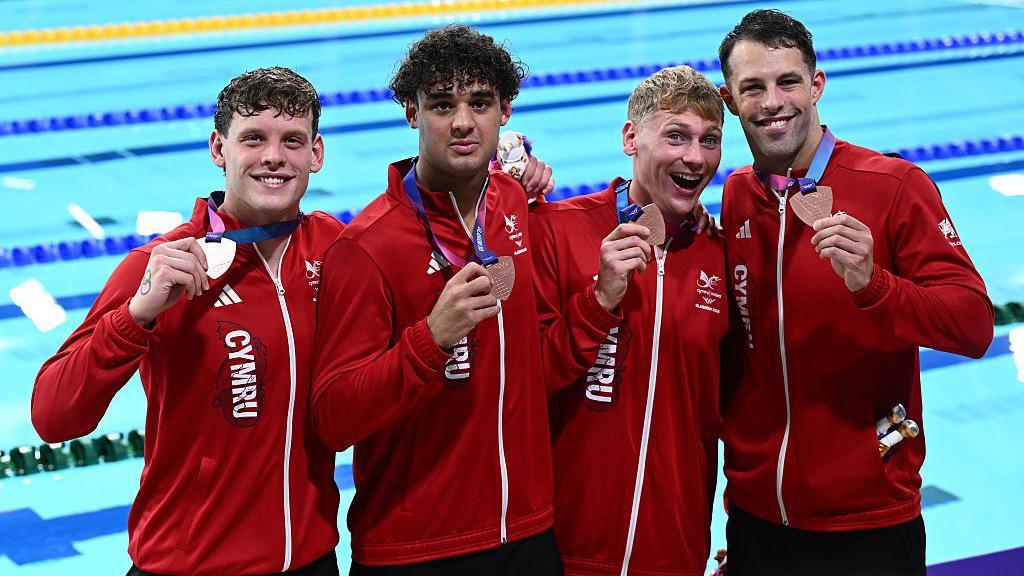 ronze medalists Matt Richards, Dan Jones, Kieran Bird and Tyler Melbourne-Smith of Team Wales pose for a photo. They are standing in front the pool wearing their Team Wales red jackets and bronze medals.