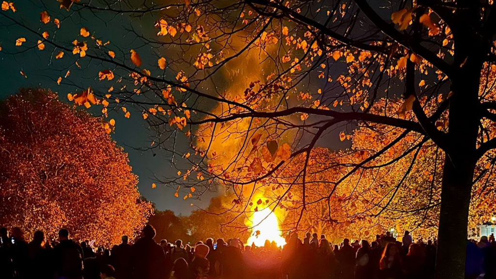 A stunning nighttime shot of a bonfire night with crowds of people and beautiful autumnal-coloured leaves on the trees, shining in the bonfire light.